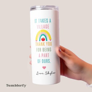 May include: White tumbler with a clear lid featuring the text "IT TAKES A VILLAGE" and "THANK YOU FOR BEING A PART OF OURS." A rainbow, apple, and heart graphic are included. The name "Love, Skylar" is also printed on the tumbler.