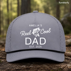 May include: Gray baseball cap with the text "AMELIA'S Reel Cool DAD EST. 2026" in white script and block letters. The cap features a black rope detail and a fish graphic. The background is a blurred outdoor scene.