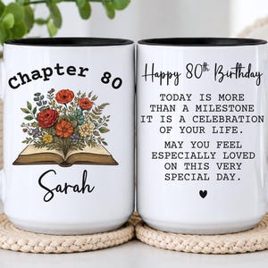 May include: Two white ceramic mugs with black handles and rims. One mug features an open book with a floral bouquet and the text Chapter 80 Sarah. The other mug displays the text Happy 80th Birthday followed by a birthday message and a small heart.