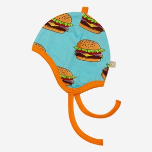 Op de afbeelding: Een baby muts met een lichtblauwe achtergrond en een herhaald patroon van cartoon hamburgers. De muts heeft een oranje rand en strikjes. De hamburgers hebben bruine broodjes, sla, tomaat, ui en kaas.