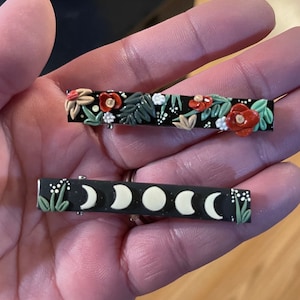 May include: Two black hair clips with decorative designs. The first clip has a floral design with red, green, and white flowers. The second clip has a moon phase design with white moons on a black background.
