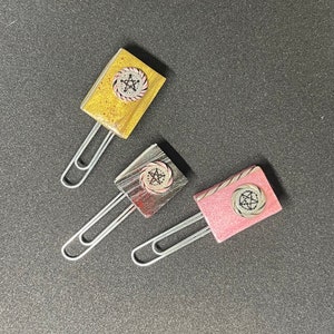 May include: Three decorative paper clips with different colored backgrounds and a silver star design in the center. The paper clips are gold, silver, and pink.