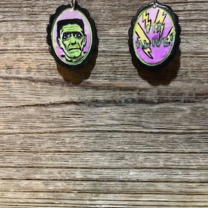 Puede incluir: Un par de pendientes con un rostro de Frankenstein negro y verde en un pendiente y las palabras "IT'S ALIVE!" en el otro pendiente. Los pendientes están sobre una superficie de madera.