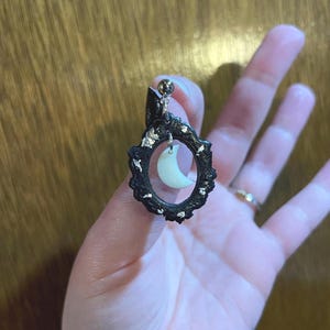 以下が含まれることがあります： 三日月型のチャームが付いた黒と銀の楕円形のイヤリング。イヤリングはテクスチャ加工された表面と銀のポストを持っています。