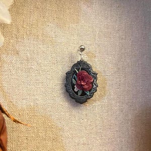 May include: A black cameo earring with a red rose and green leaves in a silver frame.