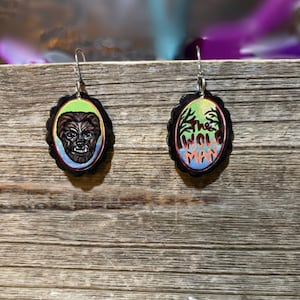 May include: A pair of earrings with a black oval-shaped design. One earring features a werewolf face with green, blue, and red colors. The other earring has a green, blue, and red background with the text "The Wolf Man" in black.