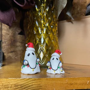 May include: Two white ghost ornaments with red Santa hats and colorful string lights wrapped around them. The ghosts are made of clay and are sitting on a wooden surface.