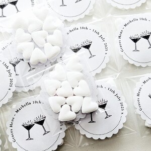 May include: Small clear plastic bags filled with white heart-shaped candies. Each bag is sealed and placed next to white scalloped-edge circles with a martini glass design and text that reads "Graces Hens - Marbella 10th - 13th July 2025".