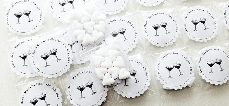 May include: Small clear plastic bags filled with white heart-shaped candies. Each bag is sealed and placed next to white scalloped-edge circles with a martini glass design and text that reads "Graces Hens - Marbella 10th - 13th July 2025".