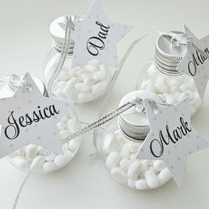 May include: Four small clear glass jars filled with white candy, each with a silver lid and a star-shaped tag with a name written on it. The names are Jessica, Dad, Mark, and Mia.