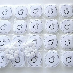May include: Small, clear plastic bags containing white heart-shaped candies and round white favors. Each favor has a diamond ring graphic and the text "LISA'S HEN MARBELLA OCTOBER 2025" printed in black. The favors have a scalloped edge.