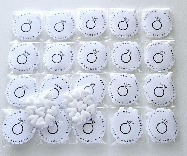 May include: Small, clear plastic bags containing white heart-shaped candies and round white favors. Each favor has a diamond ring graphic and the text "LISA'S HEN MARBELLA OCTOBER 2025" printed in black. The favors have a scalloped edge.