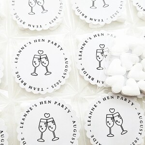 May include: White, scalloped-edge party favors with a champagne glass design and text that reads "LEANNE'S HEN PARTY AUGUST 2025 TEAM BRIDE". Some favors are packaged with heart-shaped candies.