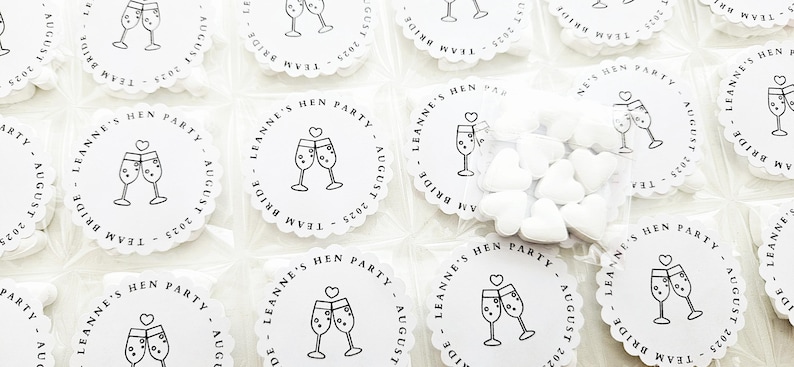 May include: White, scalloped-edge party favors with a champagne glass design and text that reads "LEANNE'S HEN PARTY AUGUST 2025 TEAM BRIDE". Some favors are packaged with heart-shaped candies.