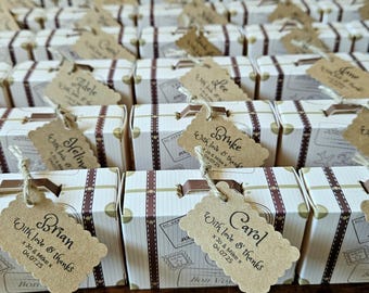 May include: Rows of miniature suitcase-shaped favor boxes with brown accents and decorative tags. Each box has a tag with a name and the text "With love & thanks" and a date. The boxes are arranged in neat rows.