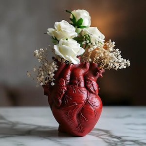 May include: A red, heart-shaped vase filled with white roses and small, dried white flowers. The vase has detailed anatomical features and sits on a marble surface. The overall theme is a unique floral arrangement.
