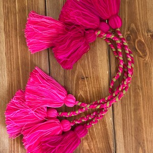 May include: Five pink and fuchsia tassels with a green and gold braided cord. The tassels are made of yarn and have a fringe at the bottom.