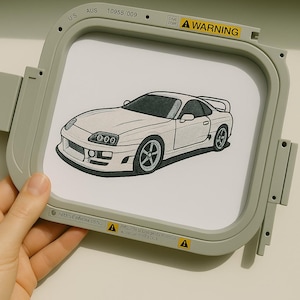 以下が含まれることがあります： 白いスポーツカーの刺繍デザイン。黒いホイールとスポイラーを備えた車のイラストが、グレーの刺繍枠に収められています。「WARNING」の文字が枠に表示されています。刺繍用デザインです。