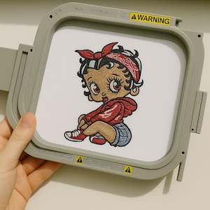 May include: Embroidered design of a cartoon baby girl wearing a red hoodie, denim shorts, and red sneakers, with a red bandana. The design is set in a gray embroidery hoop. The hoop has the text "U-S", "AUS", "A WARNING", and "REMOVE BEFORE USING".