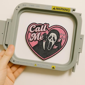 May include: Embroidered patch with a black figure, white mask, and knife, set within a pink heart, with the words "Call Me". The patch is in a gray embroidery hoop with warning labels and text.
