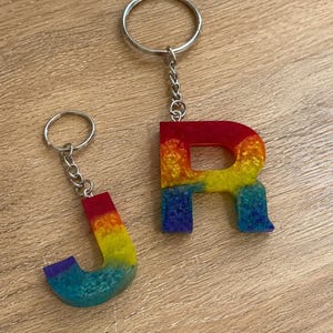 May include: Two letter-shaped keychains featuring the letters J and R. Each keychain is crafted from a multi-colored resin material with a gradient effect transitioning from red at the top to yellow, green, and blue at the bottom, attached to a silver metal ring.
