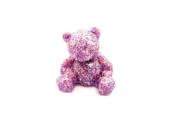 Puede incluir: Una pequeña figurita de oso de peluche rosa con purpurina. El oso está sentado con las piernas cruzadas y tiene un acabado brillante.