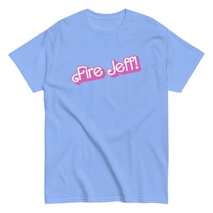 Puede incluir: Camiseta azul claro con el texto "Fire Jeff!" en rosa, contorneado en blanco. El texto, en una fuente cursiva juguetona, está centrado en la parte delantera. La camiseta es de manga corta y cuello redondo.