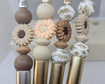 May include: A collection of decorative pens with beaded accents. The pens feature a variety of designs, including floral and patterned beads in neutral tones, and gold and silver metal barrels. The pens are held in a hand.