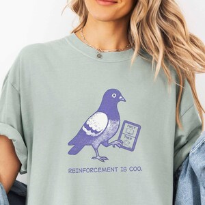 Könnte beinhalten: Ein salbeigrünes Kurzarm-T-Shirt mit einer violetten Grafik eines Taubenvogels, der ein kleines Schild mit der Aufschrift FIRST THEN hält. Unter der Taube steht der Text REINFORCEMENT IS COO. Das T-Shirt hat einen Rundhalsausschnitt und einen lockeren Schnitt.