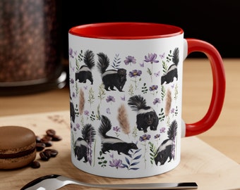 May include: White ceramic mug with a red interior and handle. The mug features a repeating pattern of black and white skunks, purple flowers, and tan grass against a white background. The mug is sitting on a wooden surface.