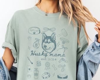 May include: A light green t-shirt with a blue husky dog illustration and the text "Husky mama est. 2024". The shirt features other dog-related icons, including a dog bowl, bone, and grooming tools.