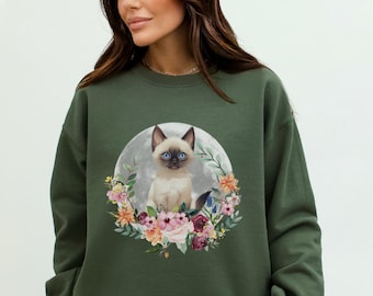 May include: A green sweatshirt with a graphic of a Siamese kitten sitting in a circle of flowers on a moon. The kitten has blue eyes and is surrounded by pink, yellow, and purple flowers.