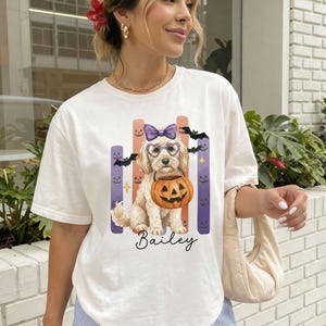 Puede incluir: Una camiseta de manga corta color marfil con un gráfico de un perro de color claro que lleva un lazo morado y gafas en forma de corazón, sosteniendo una calabaza. El diseño incluye murciélagos, rayas verticales y el nombre Bailey en cursiva.