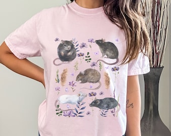 May include: A pink t-shirt with a graphic print of five rats in a floral design. The rats are black, white, and gray and are surrounded by purple flowers and green leaves.