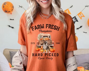 Puede incluir: Camiseta naranja con un gráfico que presenta una camioneta vintage llena de calabazas y un perro. El texto dice "Farm Fresh Pumpkins, Hand Picked, Open Daily, Est. 1980". La camiseta tiene un corte holgado.