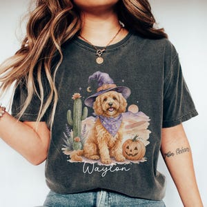 May include: A charcoal gray t-shirt featuring a graphic of a golden-colored dog wearing a purple witch hat and bandana. The design includes a cactus, a jack-o'-lantern pumpkin, and the name Waylon printed in white script below the illustration.