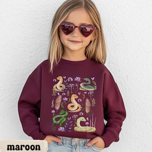 Könnte beinhalten: Ein kastanienbraunes Langarm-Sweatshirt mit einem Grafikdruck von vier gelben und grünen Schlangen, durchsetzt mit violetten Blumen, kleinen Schmetterlingen und braunen Federn. Das Sweatshirt hat einen Rundhalsausschnitt und gerippte Bündchen.