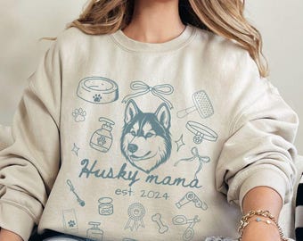 Op de afbeelding: Een licht beige sweatshirt met een blauwe husky-hond afbeelding en de tekst "Husky mama est. 2024". De afbeelding bevat verschillende hond-gerelateerde pictogrammen zoals een hondenkom, bot en borstel.