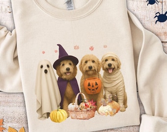 Könnte beinhalten: Cremefarbenes Sweatshirt mit Halloween-Motiv. Das Design zeigt vier Hunde in Kostümen: ein Geist, eine Hexe, ein Kürbisträger und eine Mumie. Kürbisse, ein Korb und Herbstblätter vervollständigen die Szene. Das Sweatshirt liegt auf einer Holzoberfläche.
