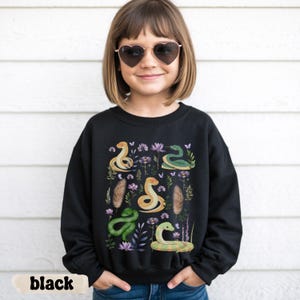 Könnte beinhalten: Ein schwarzes Langarm-Sweatshirt mit einem Grafikdruck von vier gelben und grünen Schlangen, durchsetzt mit violetten Blumen, kleinen Schmetterlingen und braunen Federbüscheln. Das Design ist mittig auf der Vorderseite des Kleidungsstücks platziert.