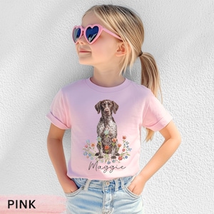 Op de afbeelding: Een lichtroze t-shirt met een afbeelding van een bruin-witte gespikkelde Duitse staande hond die tussen kleurrijke bloemen zit, met de naam Maggie in zwarte cursieve letters onder de illustratie van de hond geschreven.