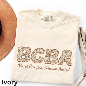 Könnte beinhalten: Elfenbeinfarbenes T-Shirt mit den Buchstaben "BCBA" in einem floralen Muster und den Worten "Board Certified Behavior Analyst" darunter. Das Shirt besteht aus Comfort Colors-Material.