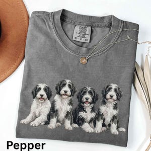 Op de afbeelding: Een antracietgrijs t-shirt met korte mouwen en een grafische print van vier zwart-witte puppy's die op een rij zitten. Het shirt heeft een ronde hals en een Comfort Colors merklabel dat aan de binnenkant van de kraag is gedrukt.