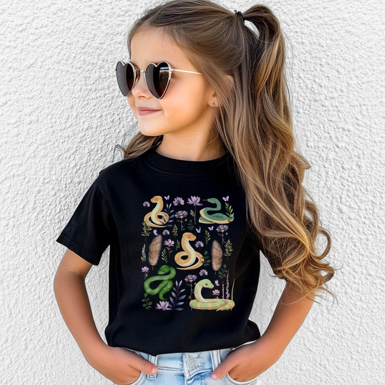 Puede incluir: Una camiseta negra con un estampado colorido de serpientes, flores y plumas. El estampado presenta una variedad de serpientes verdes y amarillas con detalles florales.