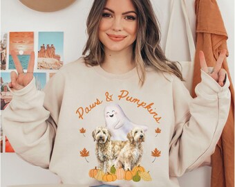 Könnte beinhalten: Sandfarbenes Sweatshirt mit dem Text "Paws & Pumpkins" und einer Grafik von zwei Hunden, einem Geist, Kürbissen und Herbstblättern. Das Wort "sand" ist unten aufgedruckt. Ein gemütliches Design zum Thema Herbst.