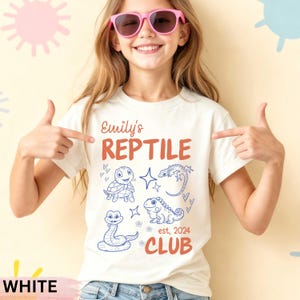May include: White t-shirt with the text "Emily's Reptile Club" and illustrations of a turtle, lizard, snake, and iguana. The text and illustrations are in red and blue. The shirt also has the text "est. 2024".