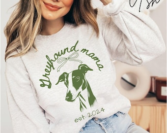 Könnte beinhalten: Ein hellgraues Sweatshirt mit dem grünen Schriftzug "Greyhound mama" und einer grün-weißen Windhund-Illustration. Der Text "est: 2024" befindet sich unter der Illustration. Das Wort "Ash" ist in Kursivschrift oben rechts geschrieben.