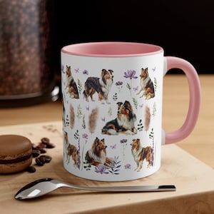 Può includere: Tazza in ceramica bianca con bordo e manico rosa. La tazza presenta un motivo ripetitivo di cani Shetland in varie pose, circondati da fiori ed erbe.