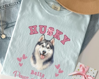 Op de afbeelding: Lichtblauw T-shirt met een roze husky-hond illustratie. De tekst "Husky" staat in roze letters boven de hond. Onder de hond staat de tekst "Bella Paws and Kisses" in roze letters.