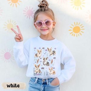 Może przedstawiać: Biała bluza z nadrukiem przedstawiającym psy corgi i fioletowe kwiaty. Słowo "white" jest wydrukowane na dole. Bluza jest noszona przez młodą osobę w różowych okularach przeciwsłonecznych, która pokazuje znak pokoju.